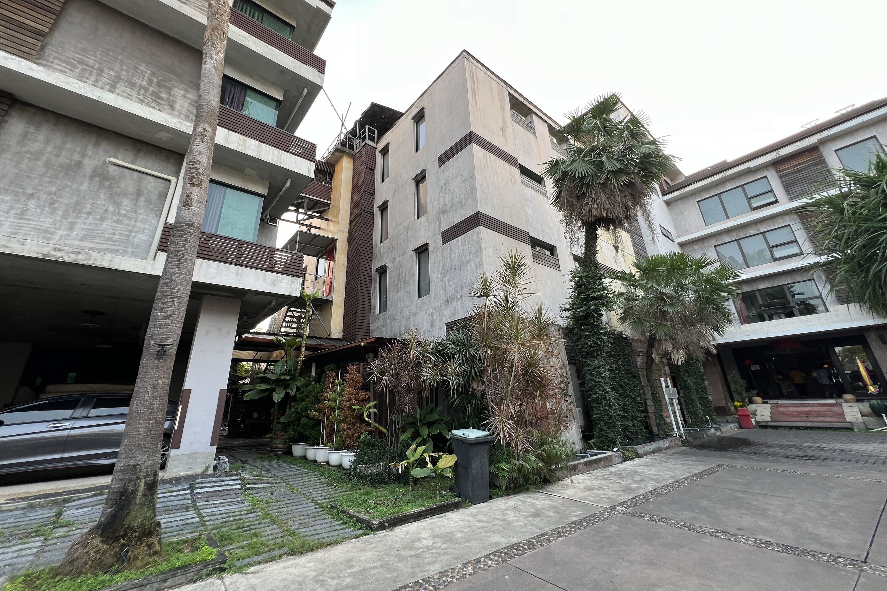 A multi-story residential building with balconies, a parking area, and a well-maintained garden with various plants.