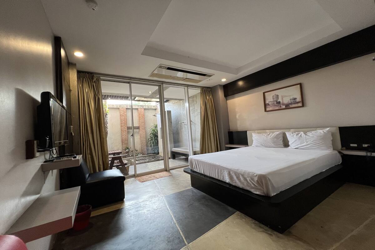 A modern hotel room with a large bed, a flat-screen TV mounted on the wall, a small seating area, and a sliding glass door leading to an outdoor area.