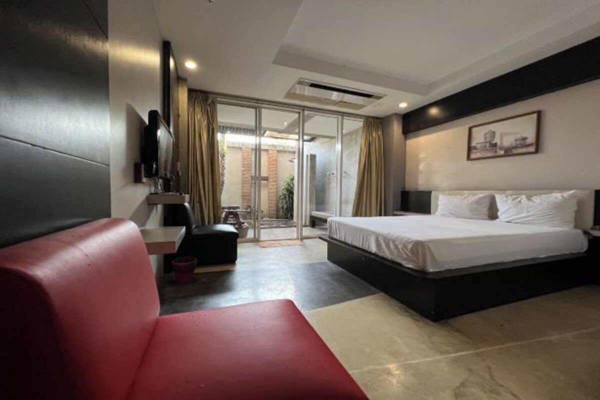 A modern hotel room with a bed, a red sofa, a small table, and a sliding glass door leading to a balcony.
