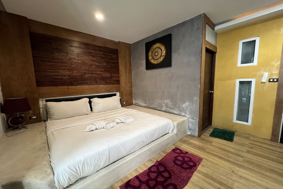 A neatly made bed with white linens and towels, a wooden headboard, a wall clock, and a small rug on the floor.