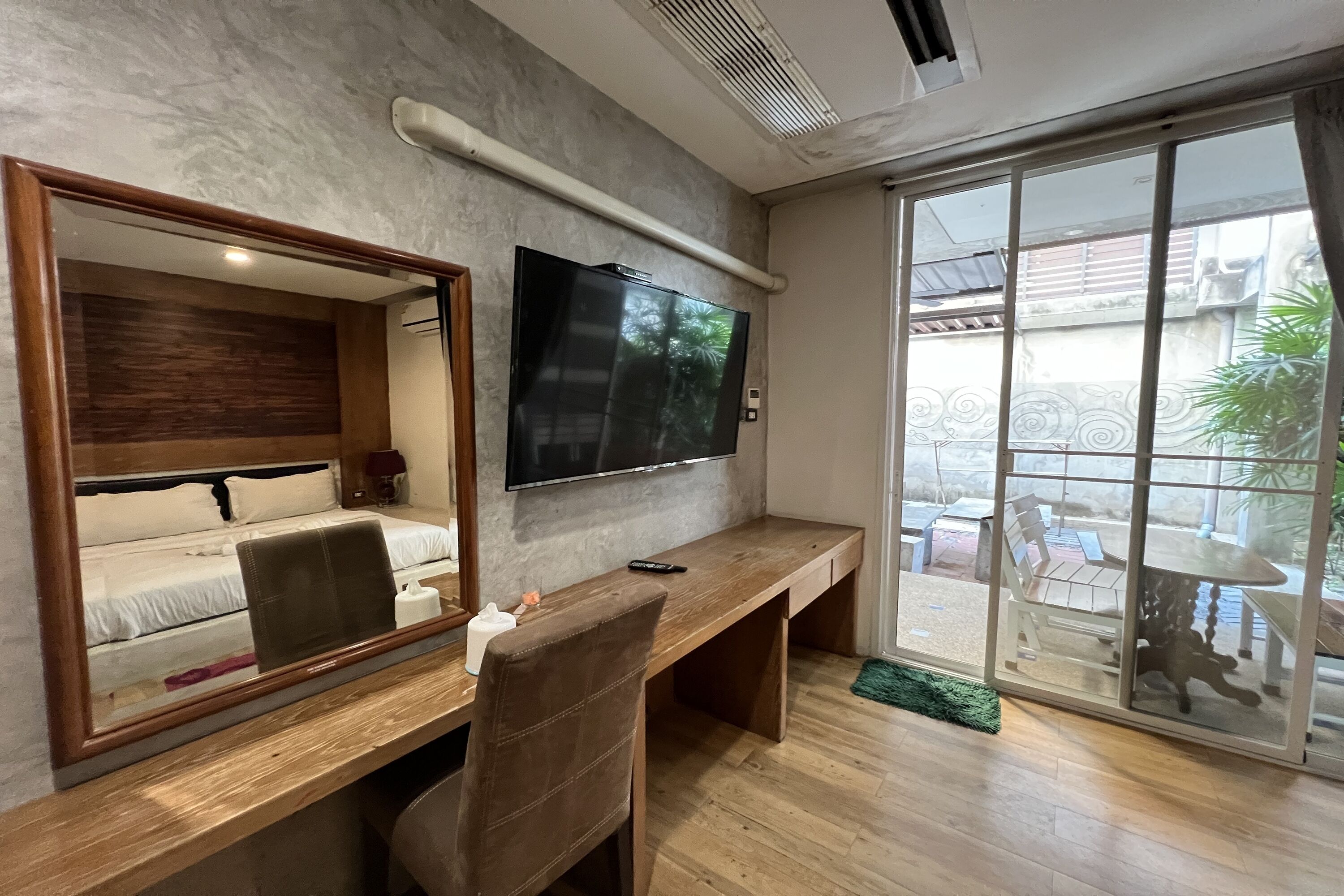 Una habitación de hotel moderna con un escritorio de madera, un espejo grande, un televisor de pantalla plana y un ventanal corredizo que da a un patio exterior.