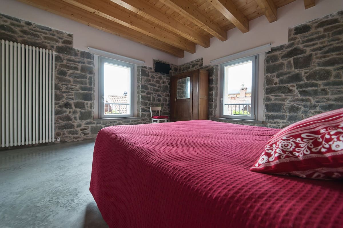 Een slaapkamer met een stenen muur, houten balken in het plafond, een rode sprei en een raam met uitzicht op gebouwen.
