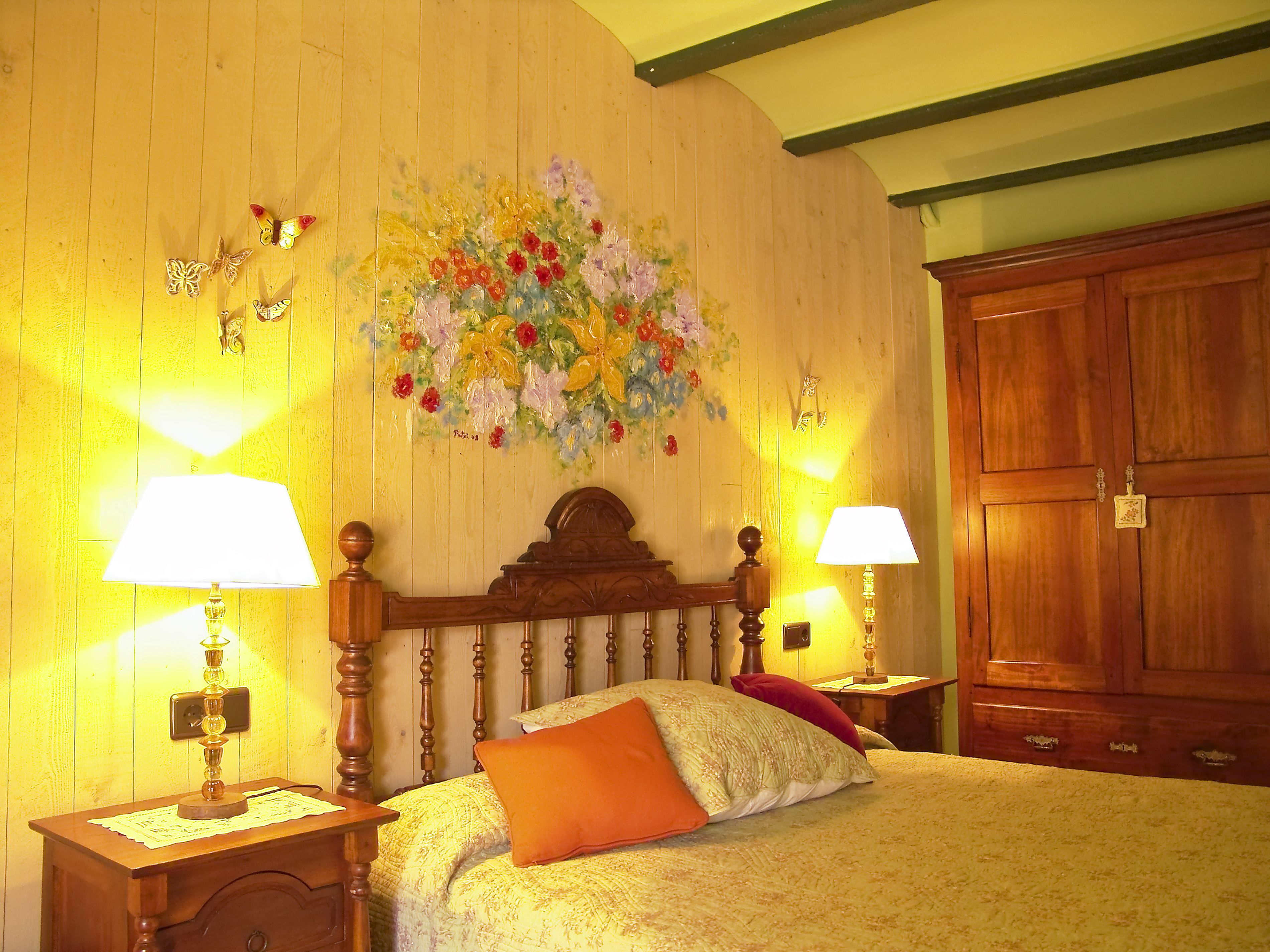 A bedroom with a wooden headboard, two bedside lamps, a floral wall decoration, and a large wardrobe.