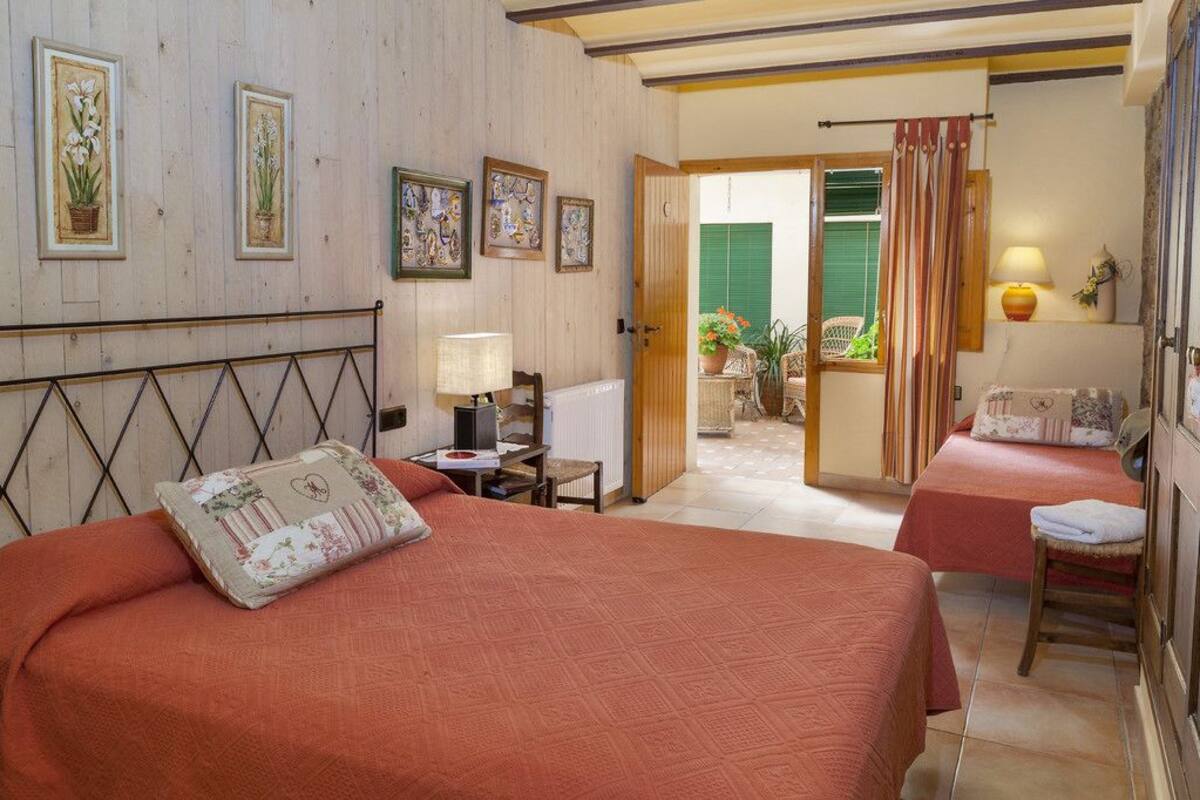 A hotel room with two beds, a desk with a lamp, and a view of the garden through an open door.