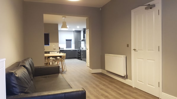 Ein modernes Wohnzimmer mit einer Couch, einem Esstisch mit Stühlen, einer Küchenzeile mit Schränken und Geräten sowie einer weißen Tür.
