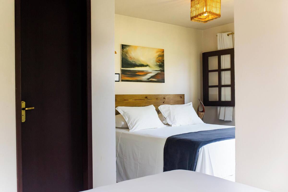 A bedroom with a wooden headboard, a bed with white bedding, a painting on the wall, and a window with curtains.