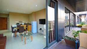 Een moderne keuken met een koelkast, magnetron en kookplaat. Er is een eethoek met een glazen tafel en stoelen, en een balkon met rieten meubels en planten.