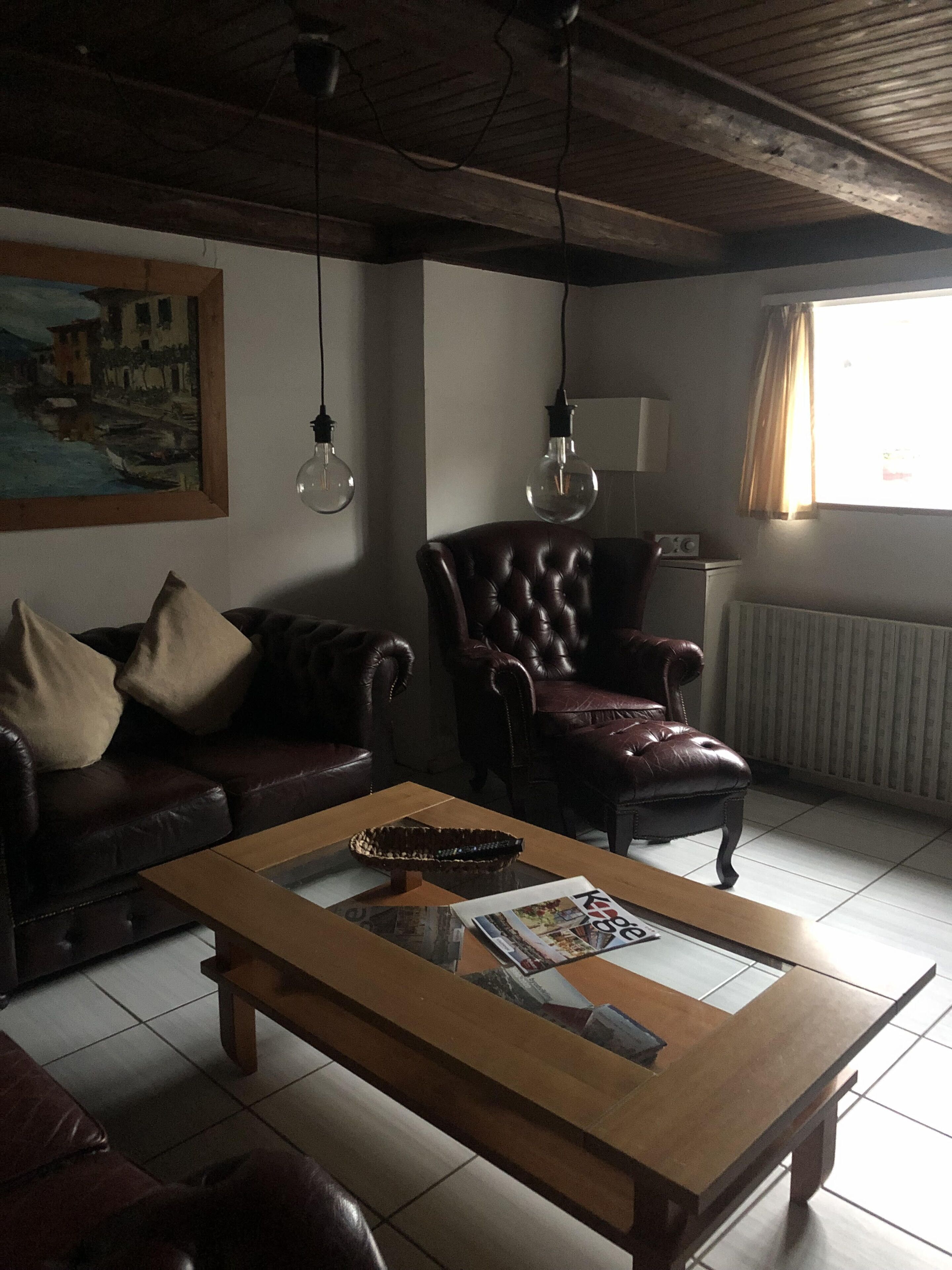 A living room with a wooden coffee table, leather sofa, and a leather armchair. There is a painting on the wall and a window with curtains.