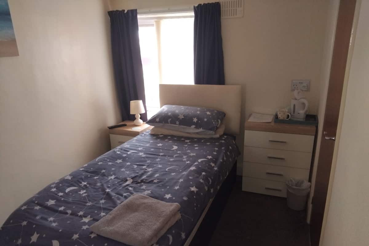 A small, neatly arranged bedroom with a bed, bedside table, lamp, and a small desk with a cup and a kettle.