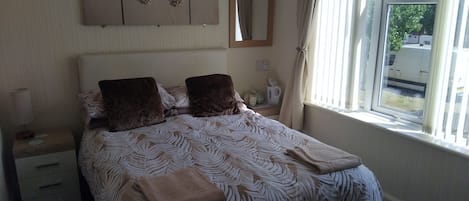 A neatly made bed with a patterned bedspread, two pillows, and a folded towel. There is a bedside table with a lamp and a small decorative item. A mirror hangs on the wall above the bed, and a window with curtains is visible.