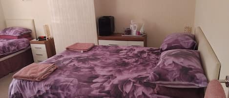 Habitación de hotel con dos camas, cada una con ropa de cama de flores moradas, una mesita de noche pequeña con una lámpara y un televisor montado en la pared.