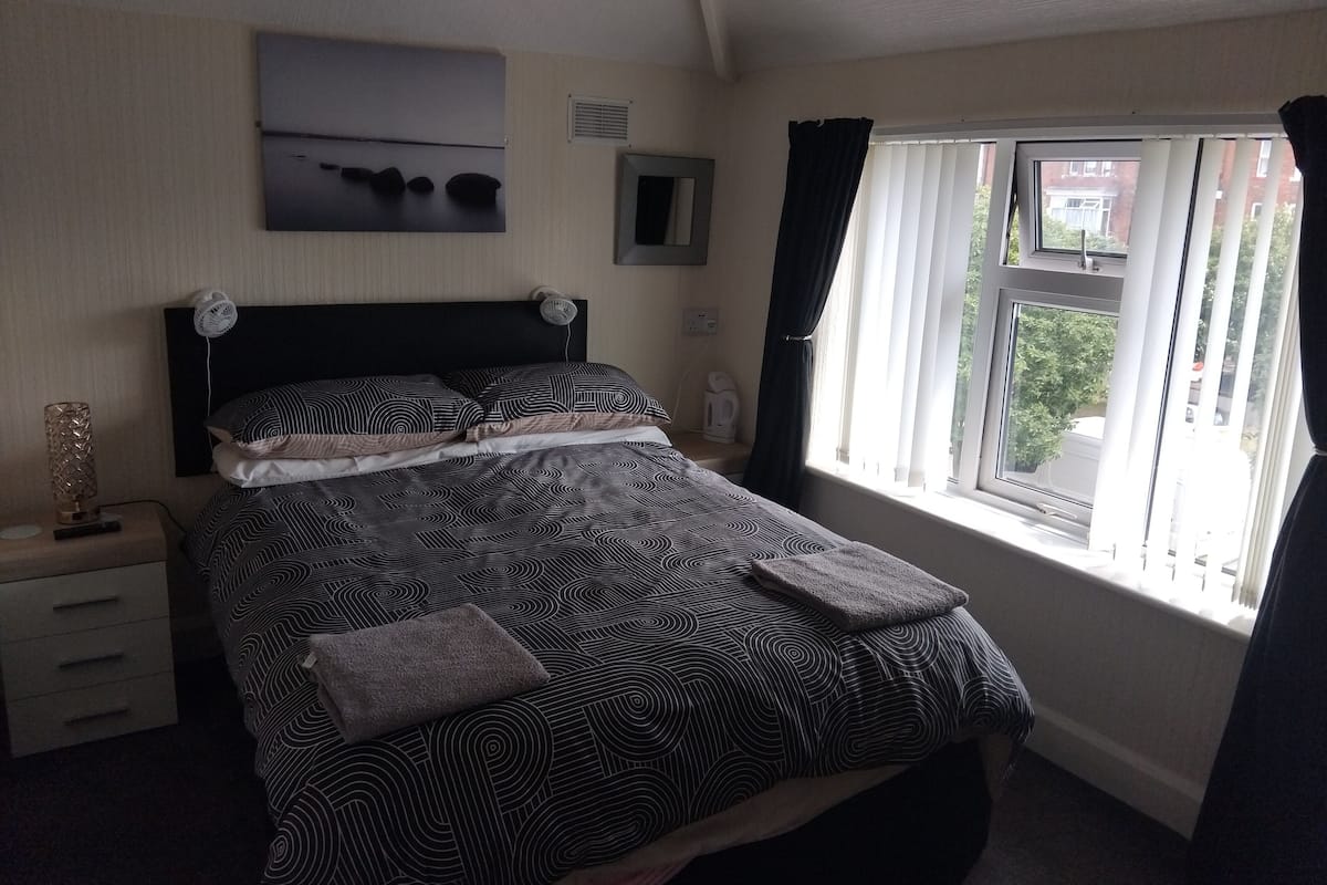 A neatly made bed with a patterned bedspread, a bedside table with a lamp, and a window with curtains.