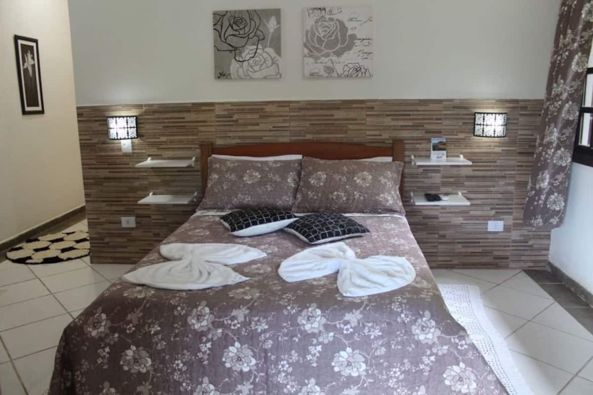 Un dormitorio con cabecera de madera, colcha estampada con motivos florales, dos mesitas de noche con lámparas y obras de arte en las paredes.