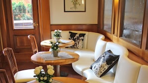 A cozy interior with a wooden door, a framed floral artwork, a white sofa with patterned cushions, and a round wooden table with a vase of flowers. - Hotel Binelli (Pinzolo)