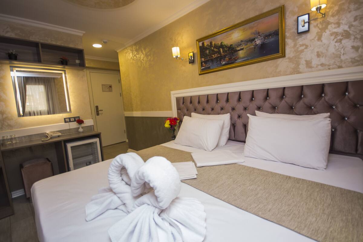 A hotel room with a large bed, a bedside table, a desk, and a chair. There is a painting on the wall and a window with curtains.