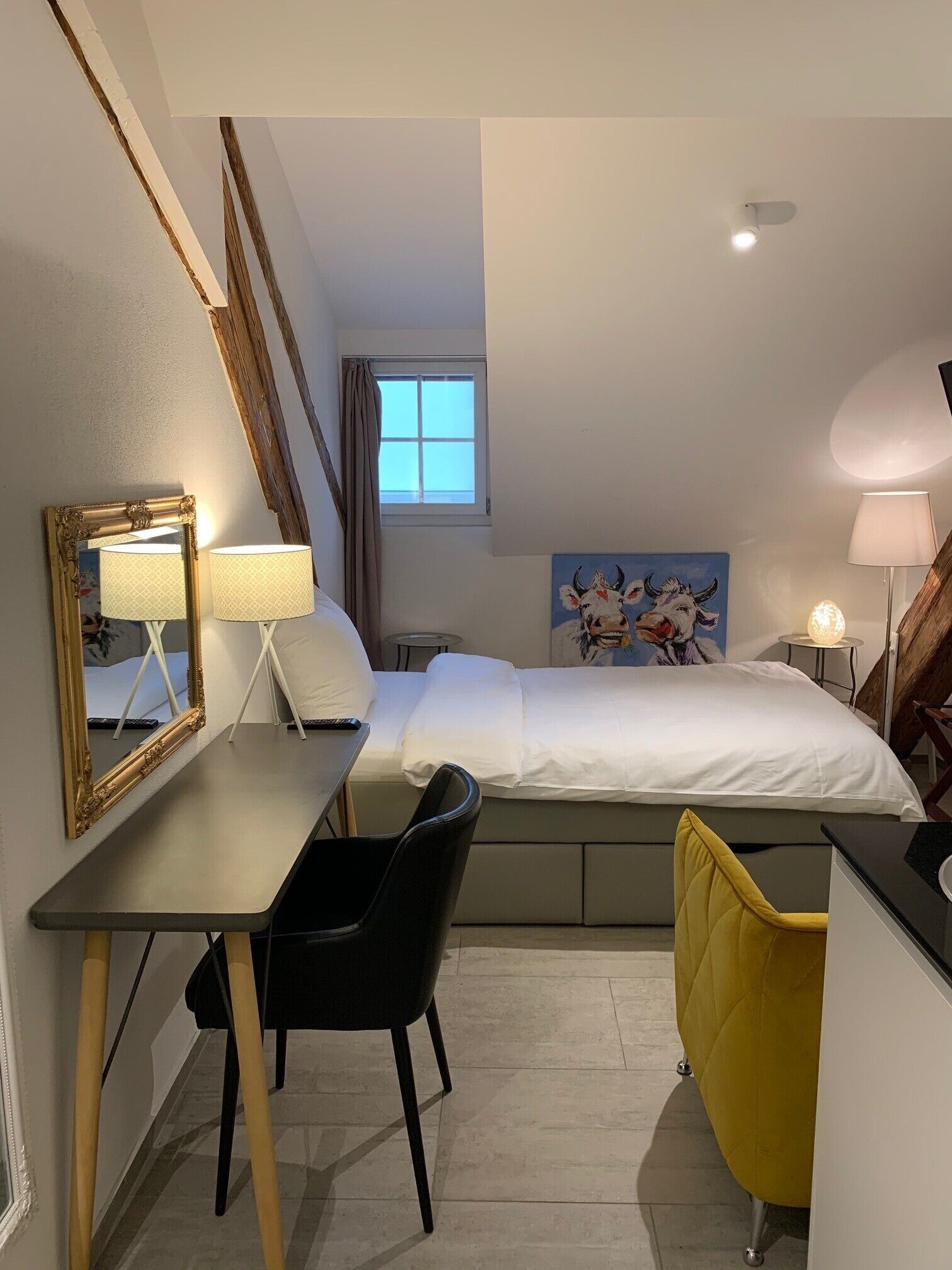 Une chambre à coucher de taille modeste, comprenant un lit, un bureau avec deux lampes, une chaise et un tableau accroché au mur.