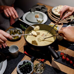 A group of people enjoying a fondue meal with various accompaniments like bread, olives, and grapes.