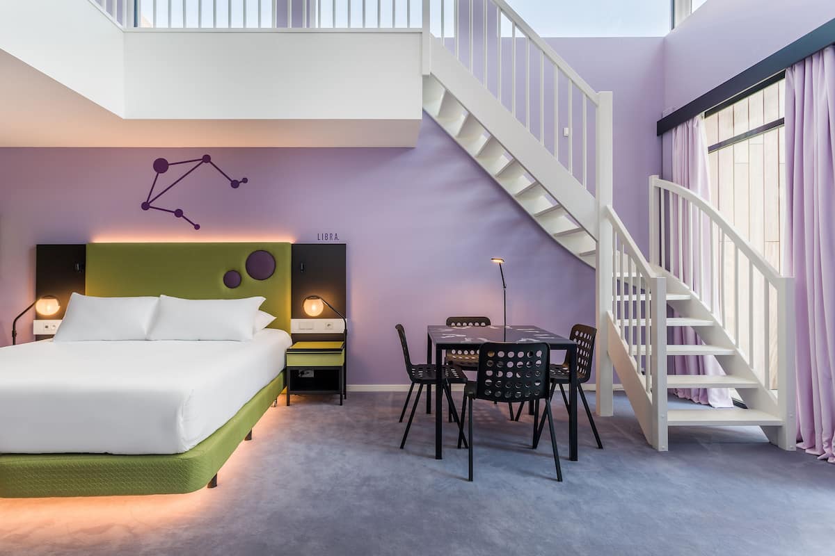 Une chambre à coucher comprenant un lit, un bureau, des chaises, un escalier et un mur décoré d’un motif représentant une constellation.