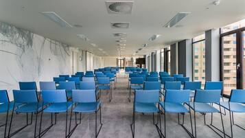 A conference room with rows of blue chairs, a grey carpet, and large windows with a view of buildings.