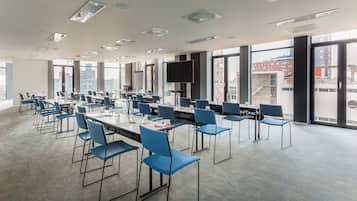 Una sala de conferencias moderna con varias mesas y sillas azules, ventanas amplias con vista a la ciudad y una televisión de pantalla plana.