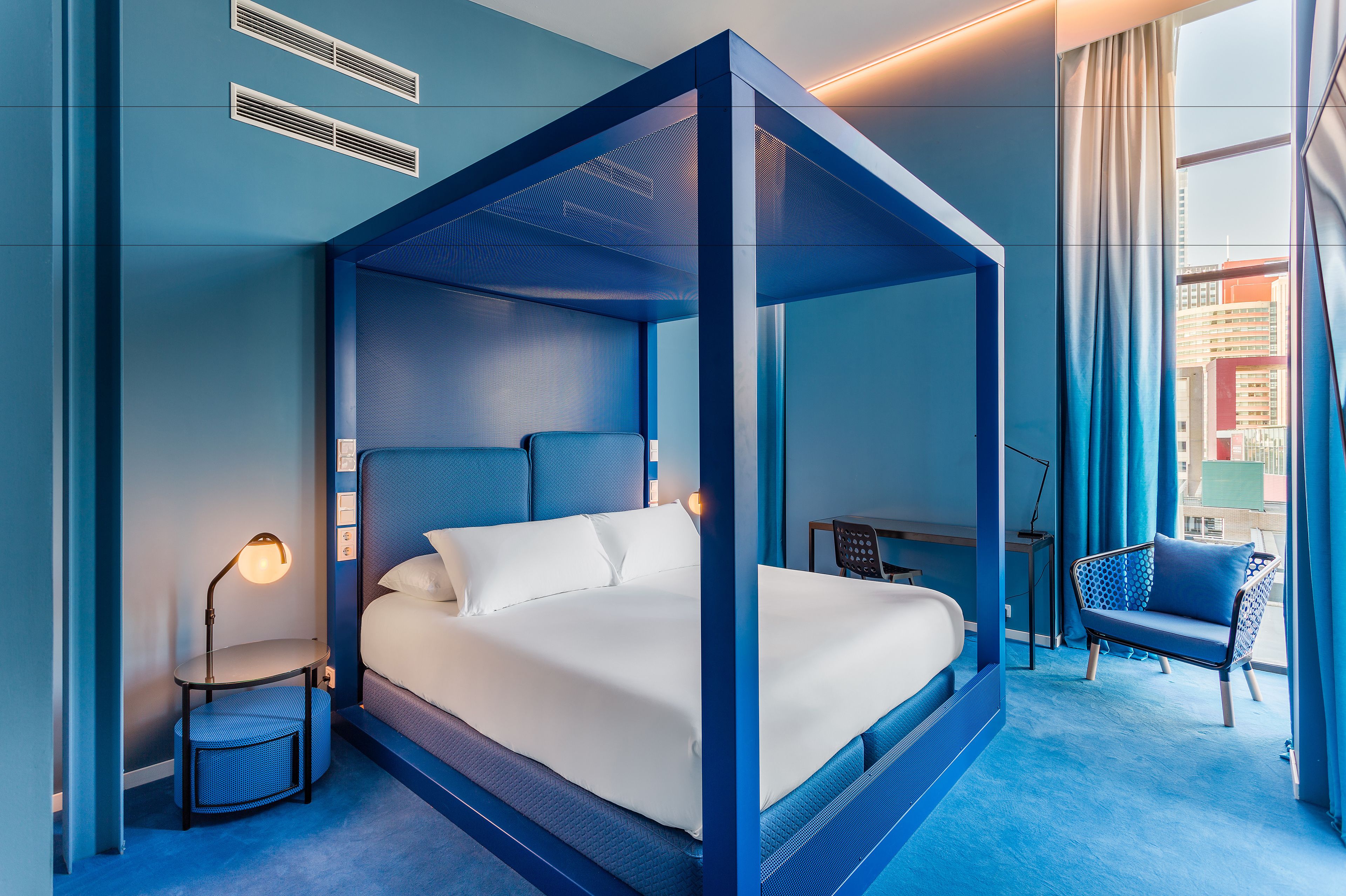 A modern hotel room with a four-poster bed, a desk with a chair, and a view of the city through large windows.