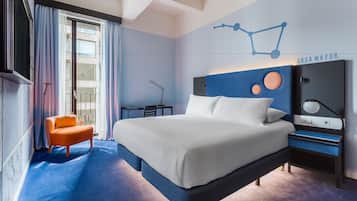 A modern hotel room with a large bed, a nightstand, a chair, and a wall-mounted TV.