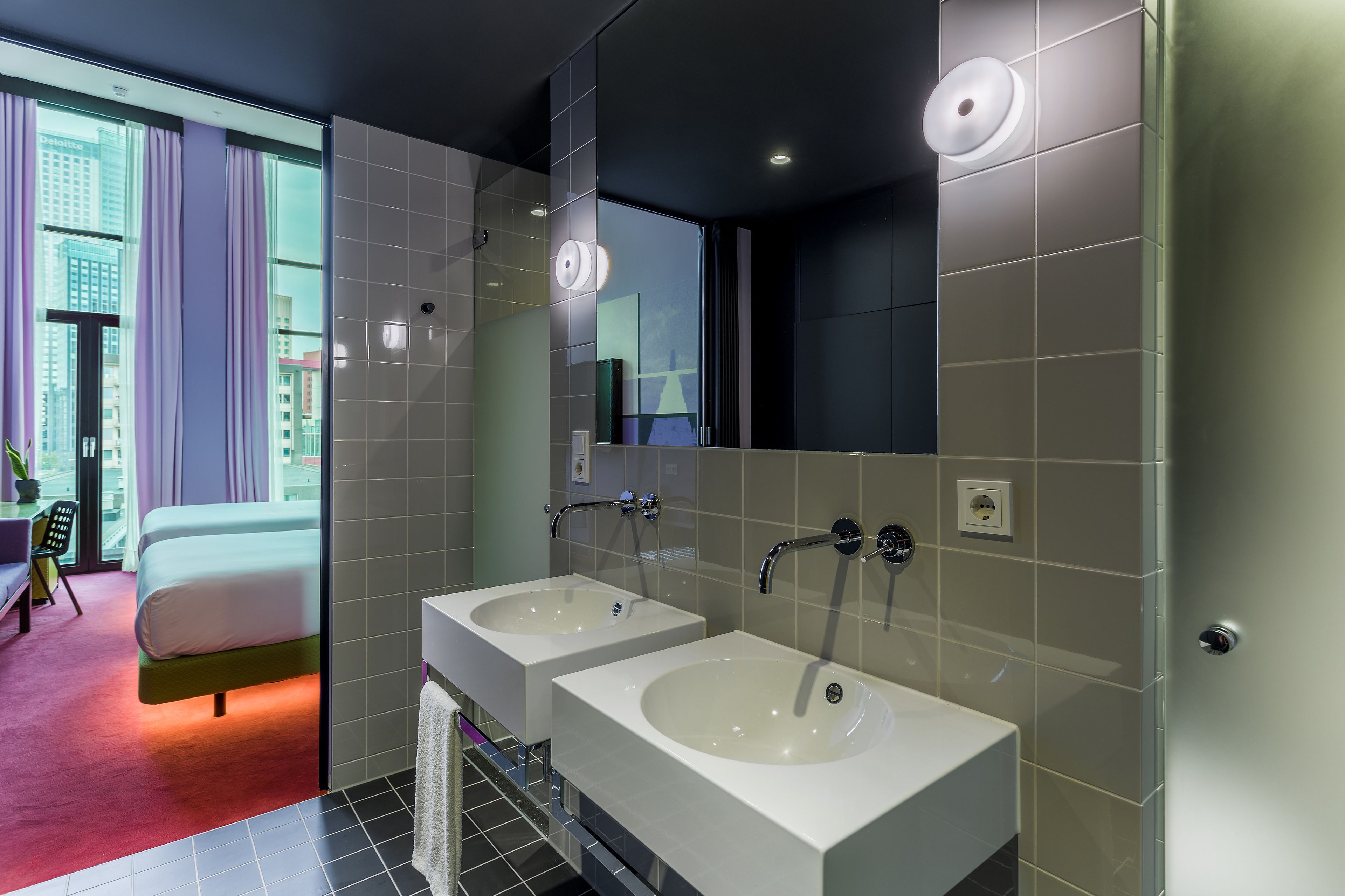 Une salle de bain moderne avec deux lavabos, un grand miroir et une vue sur le paysage urbain, visible à travers une porte en verre.