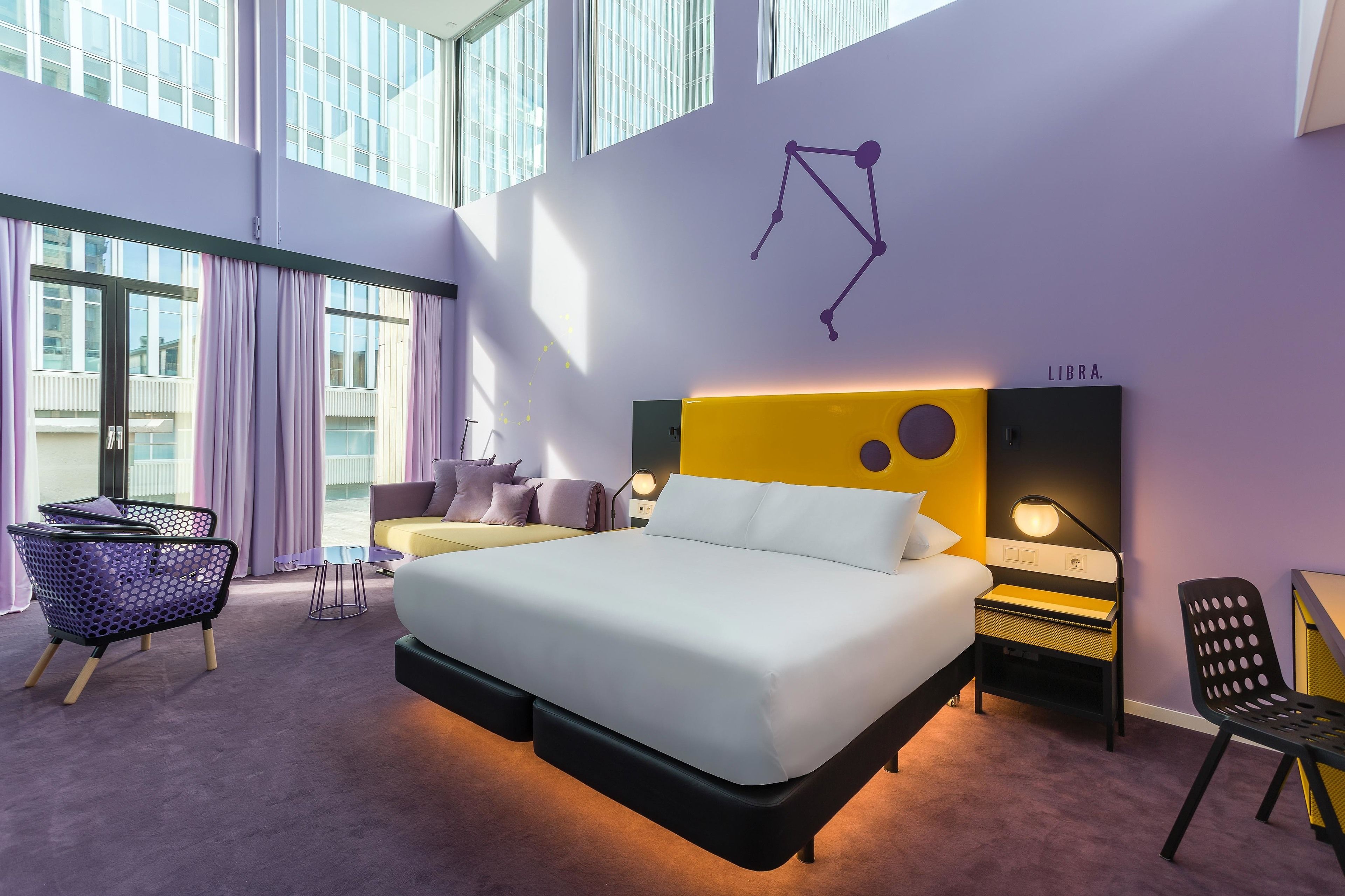 A modern hotel room with a large bed, a desk, and a chair. There is a window with curtains, a wall-mounted lamp, and a decorative wall piece.