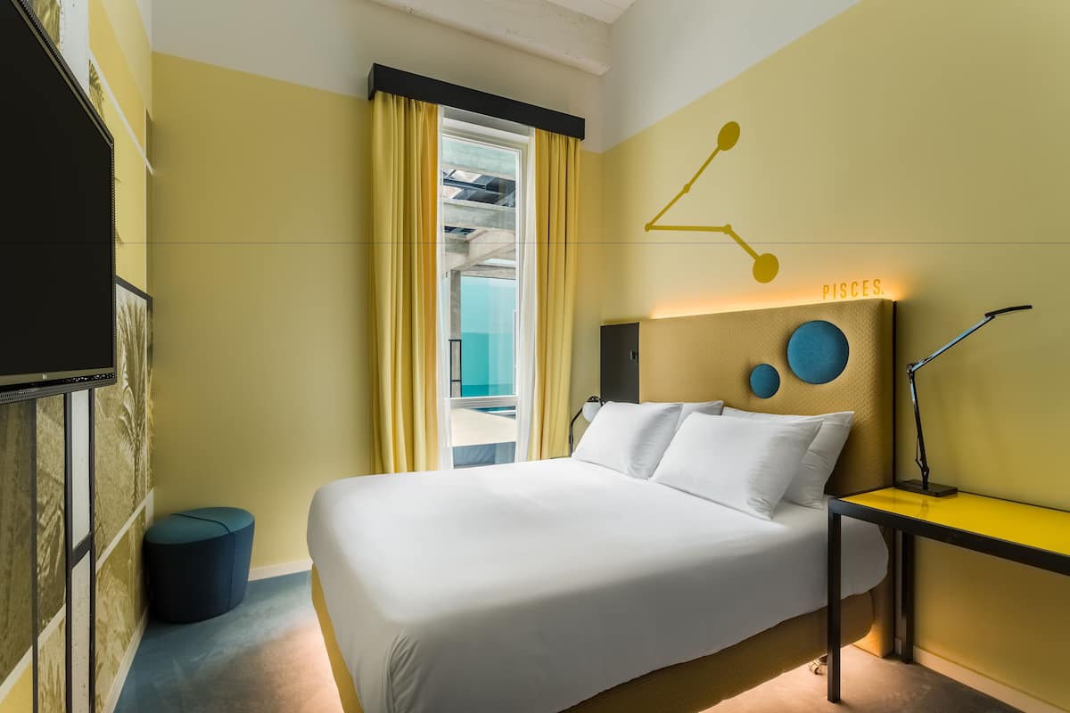 Une chambre d’hôtel avec un lit, une table de chevet, une lampe, une horloge murale et une fenêtre avec des rideaux.