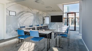 A modern conference room with a long table, blue chairs, a projector screen, and a city view through large windows.