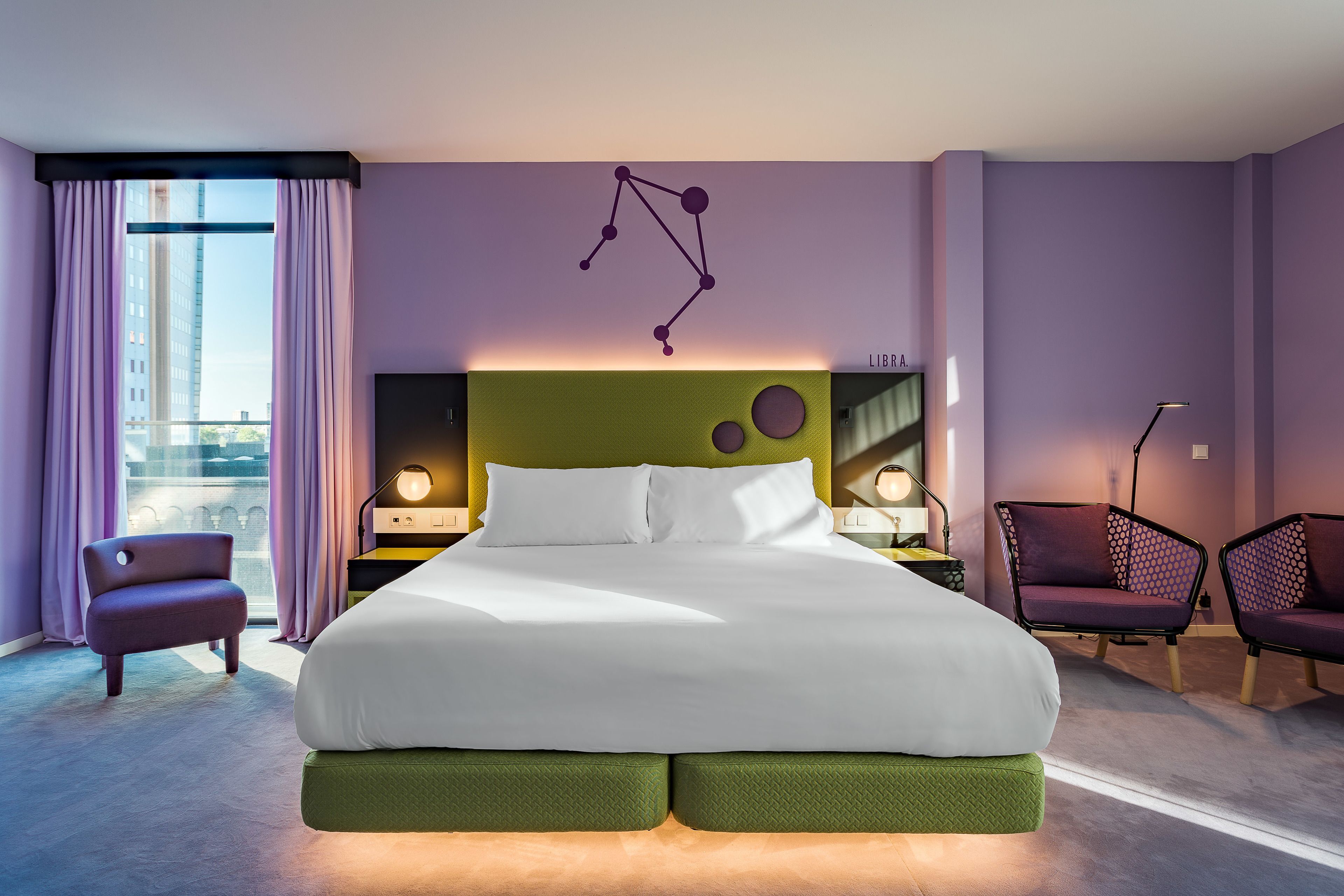 Une chambre d’hôtel moderne avec un grand lit, une vue sur la ville et des murs violets.