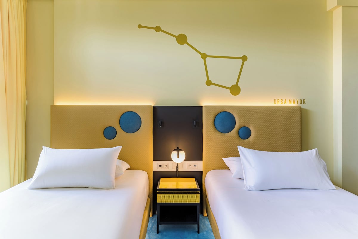 Une chambre d’hôtel avec deux lits, une table de chevet, une lampe fixée au mur et une structure moléculaire décorative.