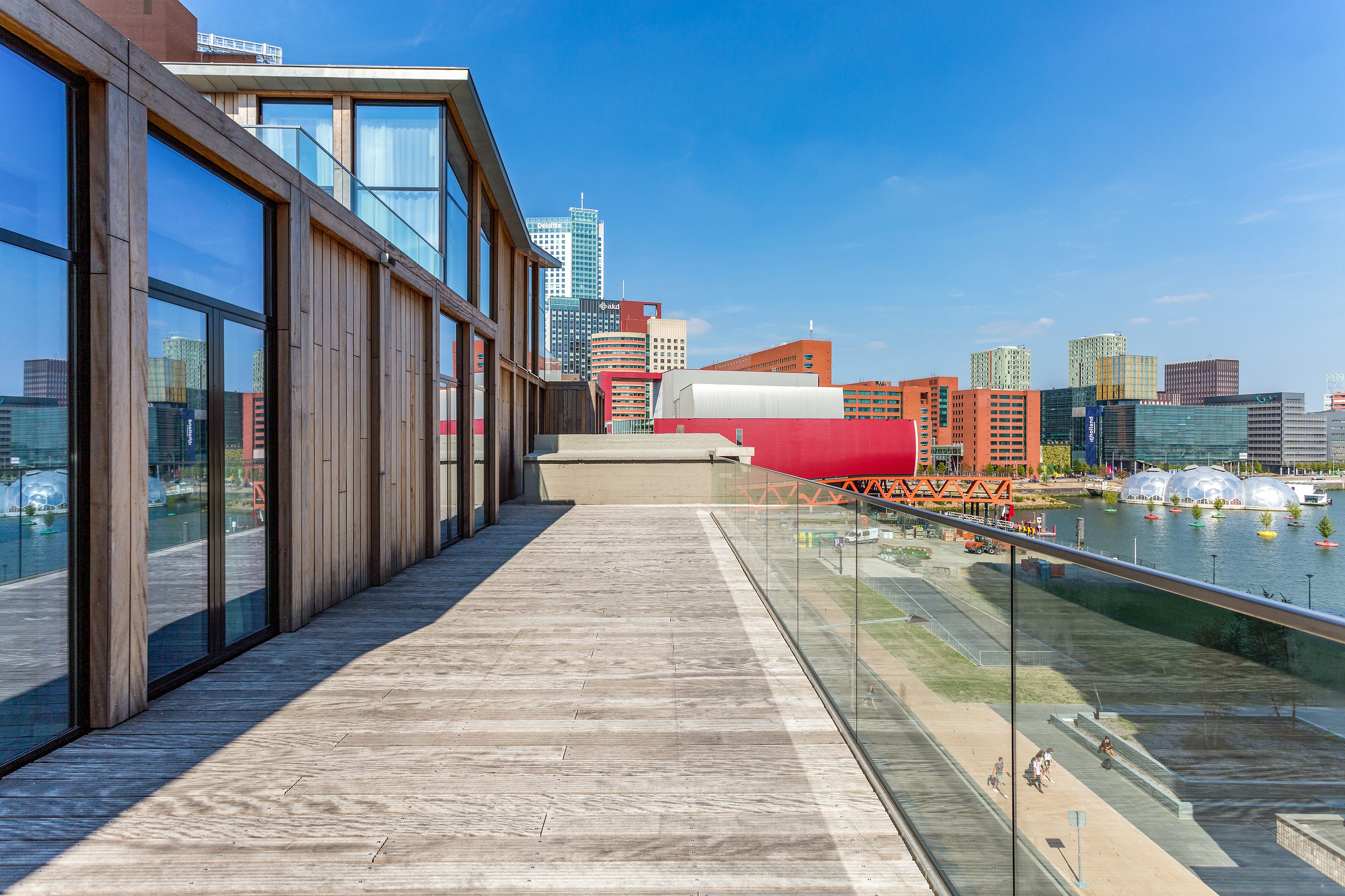 A modern building with large windows and a wooden deck overlooking a river with boats.
