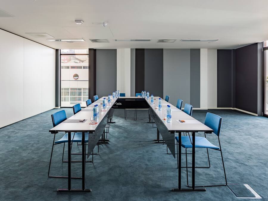 Sala de conferência com uma mesa comprida, cadeiras azuis e garrafas de água.