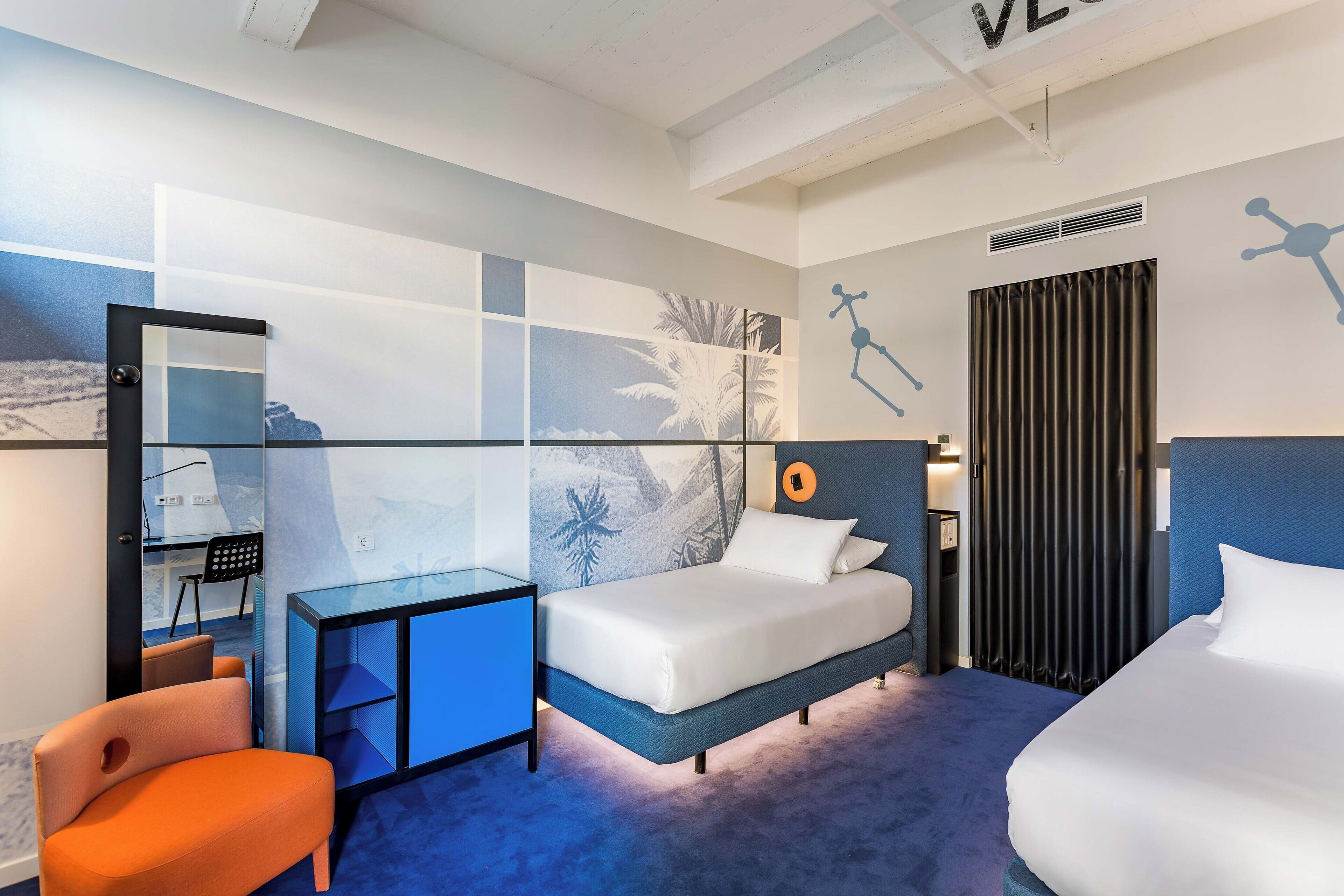 A hotel room with a bed, a desk, a chair, a mirror, and a wall with a tropical mural.