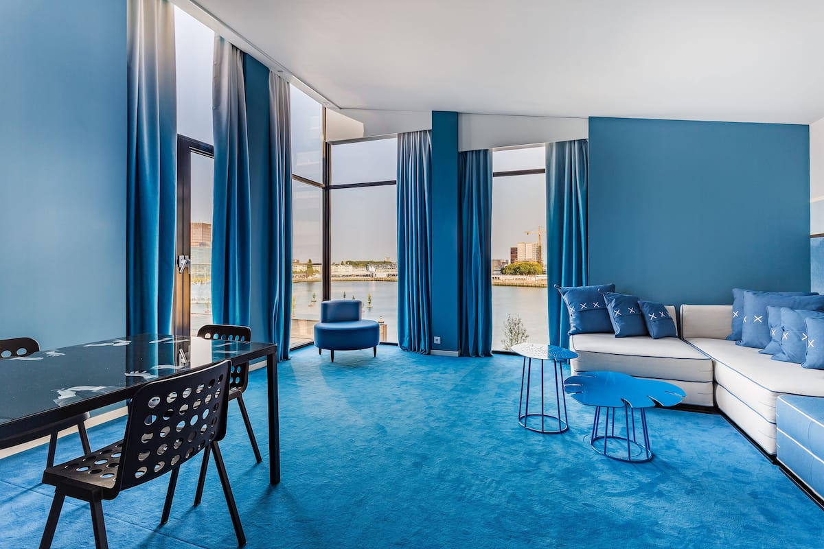 Une pièce moderne avec une table en verre, des chaises bleues, un canapé et une vue sur une étendue d’eau.