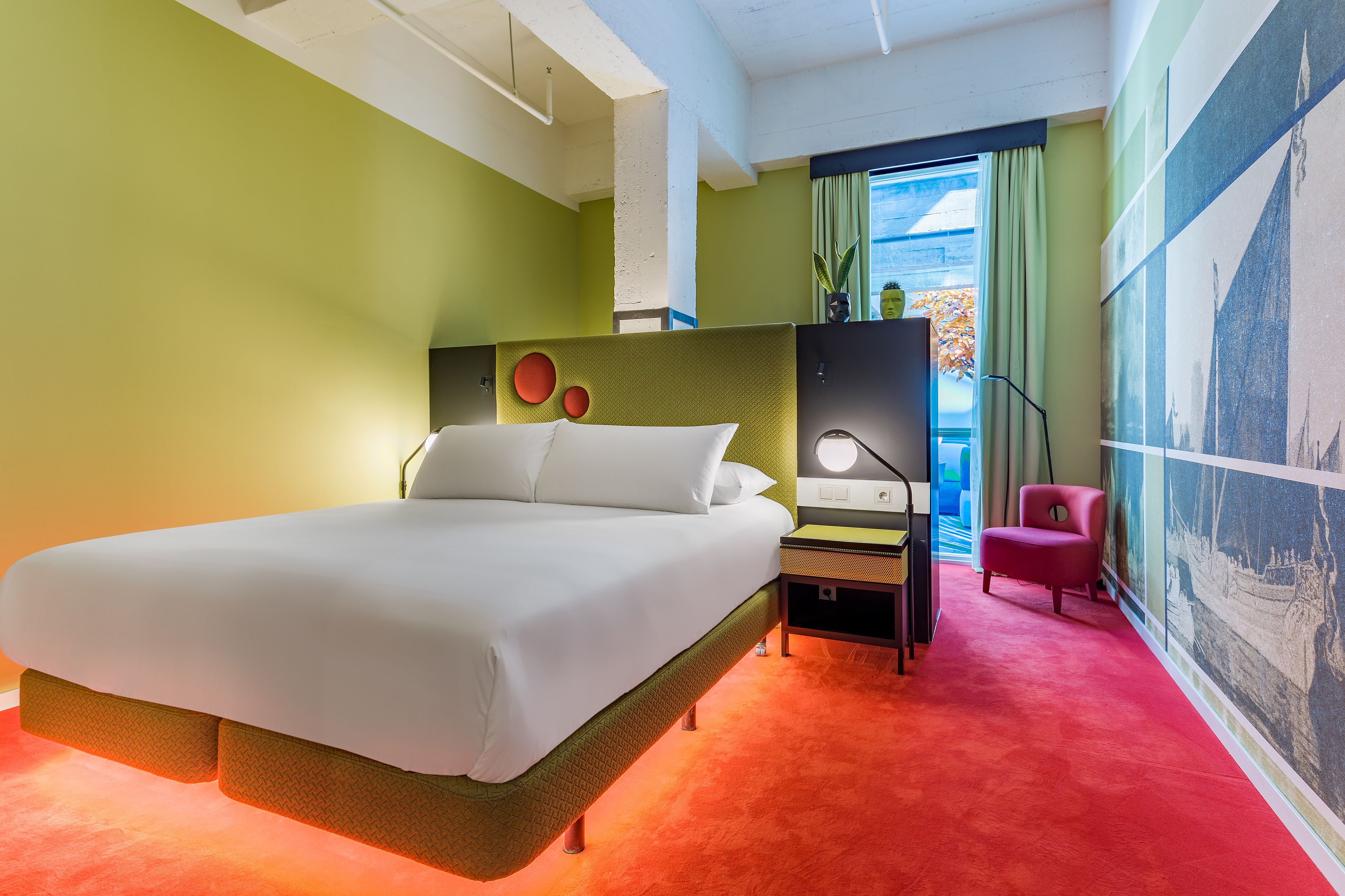 A modern hotel room with a large bed, a nightstand, a red chair, and a large window with a view.