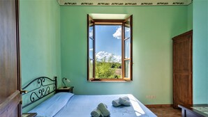 A bedroom with a bed, a nightstand, a wooden wardrobe, and a window with a view of greenery. - Bazzani Apartments (Casole d'Elsa)