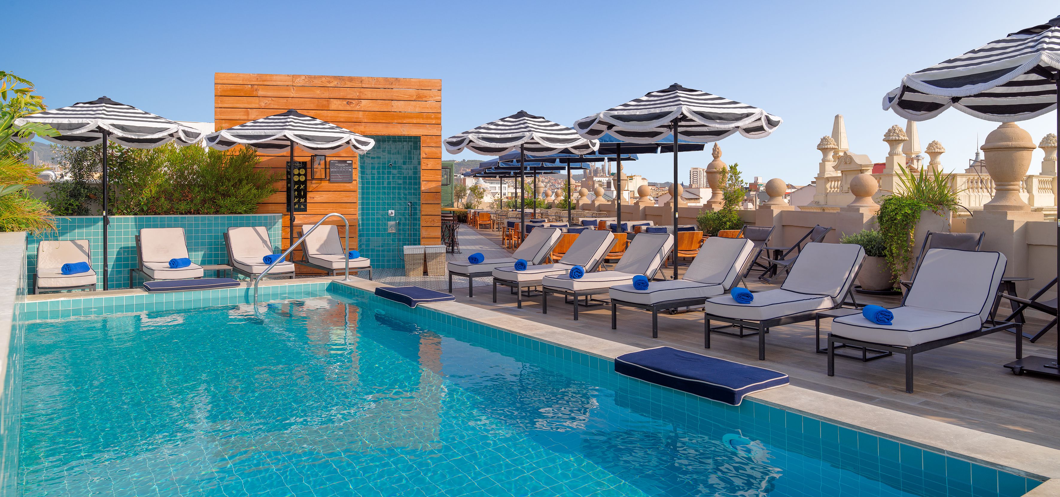 A rooftop pool area with lounge chairs and umbrellas, a wooden wall, and a view of the cityscape.