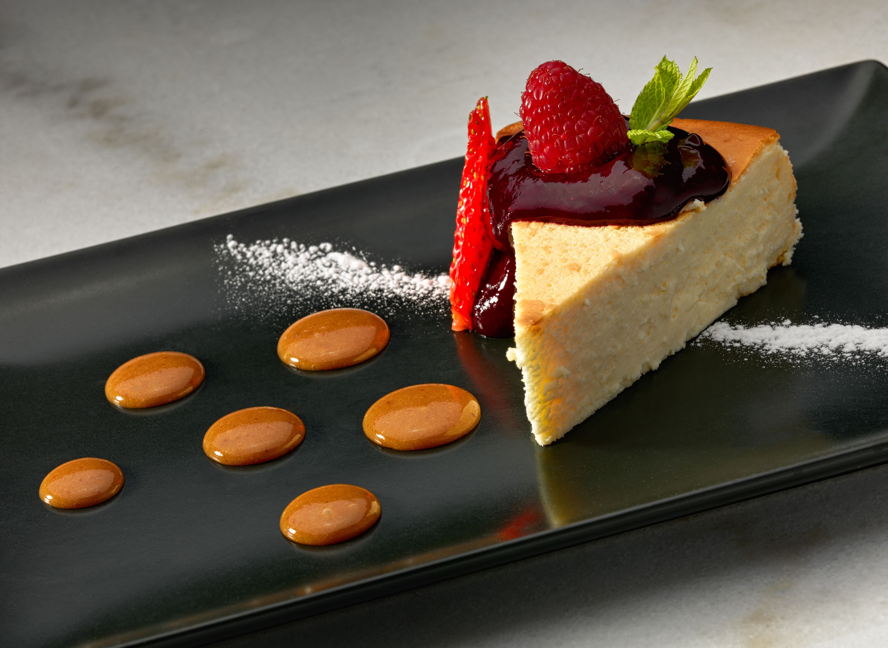 A slice of cheesecake with a berry compote and a mint leaf on a black plate.