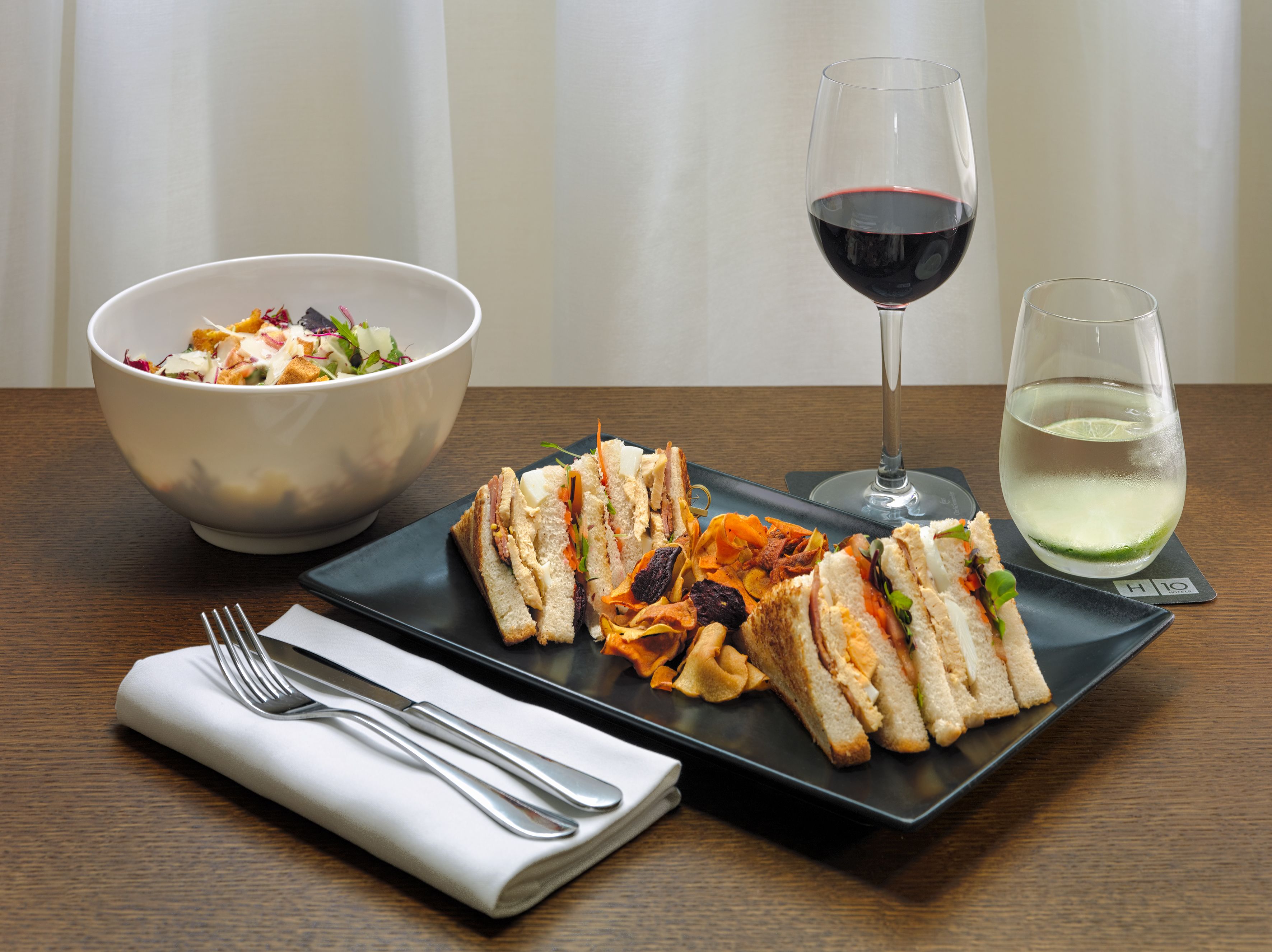 Uma mesa de jantar com um prato de sanduíches, uma saladeira, uma taça de vinho tinto e um copo de água.