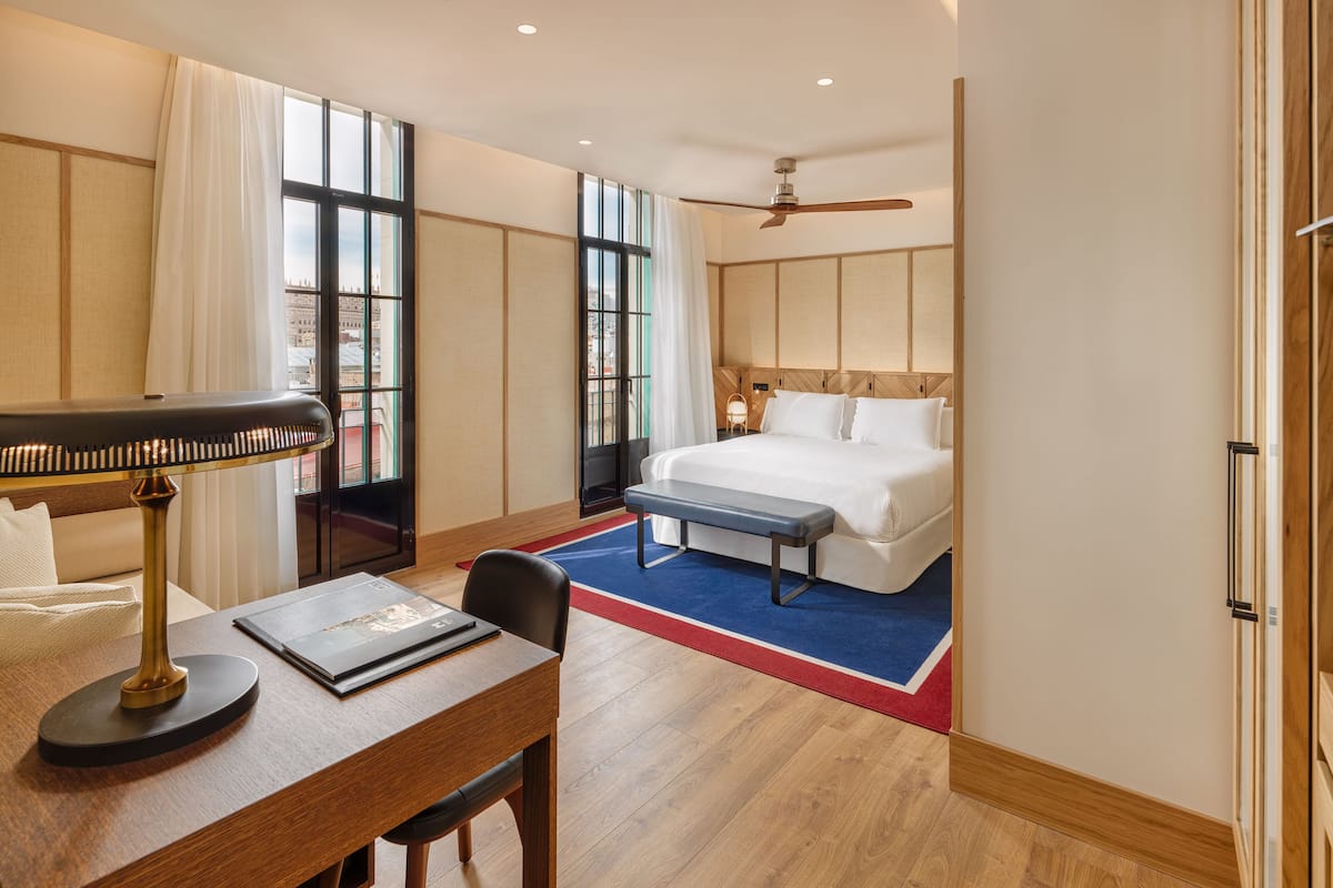 Ein Hotelzimmer mit Bett, Schreibtisch mit Lampe, einem Sessel und Blick auf die Stadt durch große Fenster.