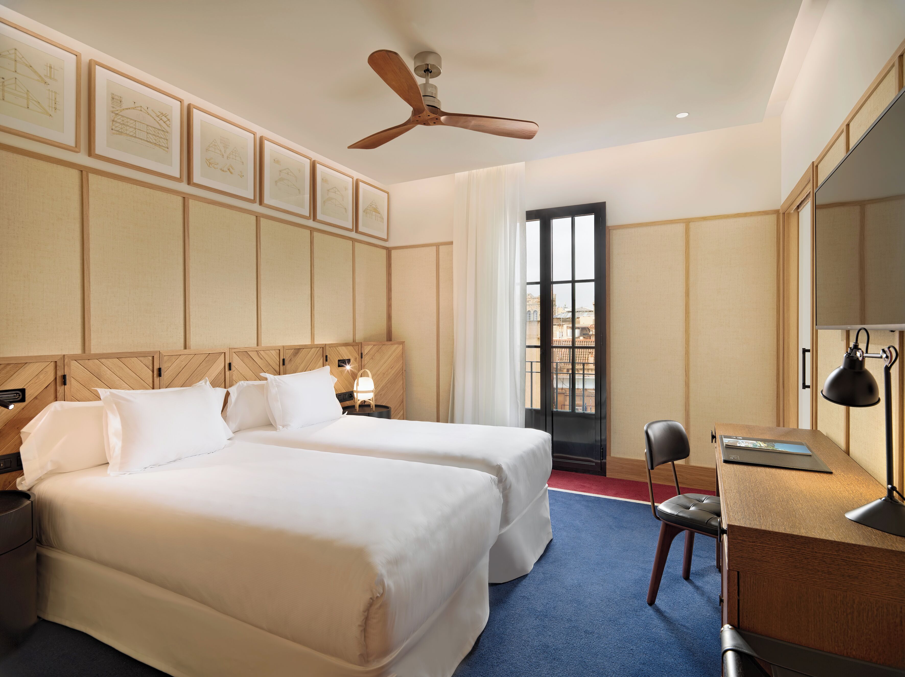 A hotel room with two beds, a desk, a chair, a ceiling fan, and framed architectural drawings on the wall.