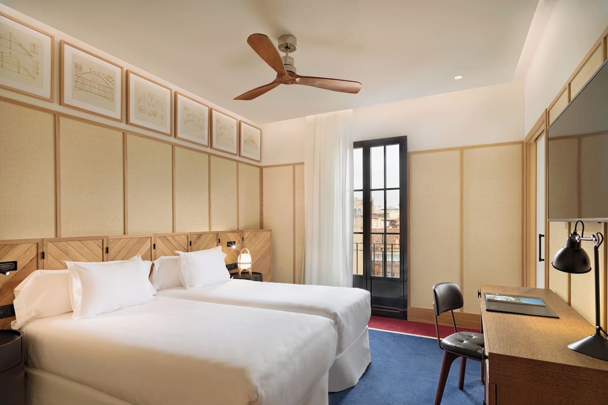 Ein Hotelzimmer mit zwei Betten, einem Schreibtisch mit Stuhl, einem Deckenventilator und gerahmten architektonischen Zeichnungen an der Wand.