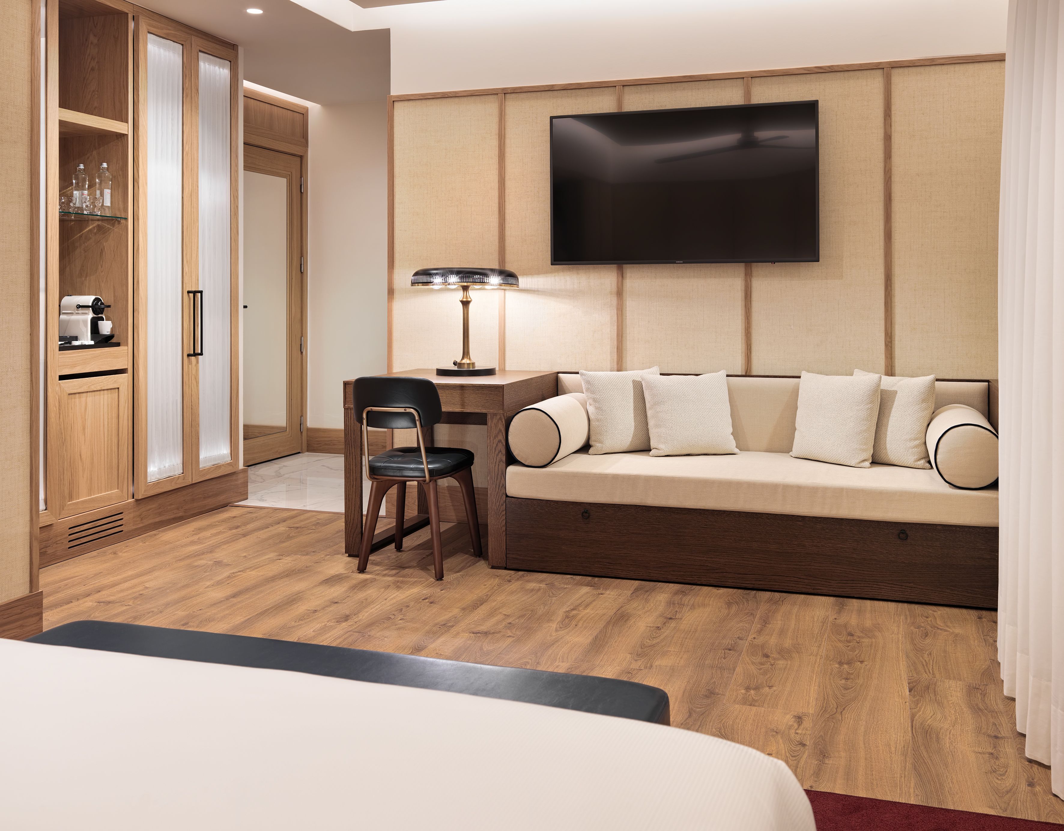 Habitación de hotel con un armario de madera, un escritorio con silla, un sofá con cojines y un televisor de pantalla plana montado en la pared.
