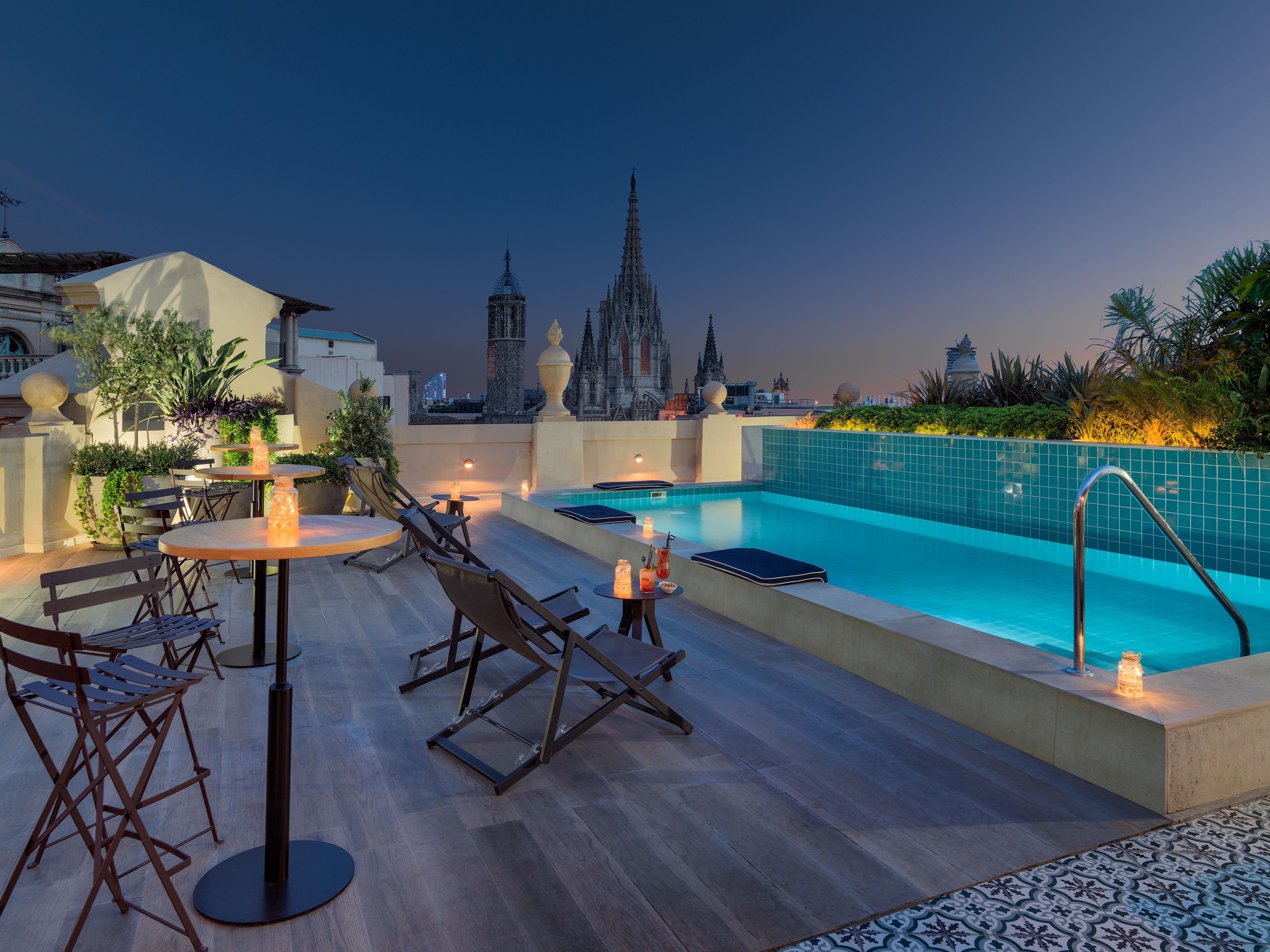 A rooftop pool area with lounge chairs and tables, a swimming pool, and a view of historic buildings in the background.
