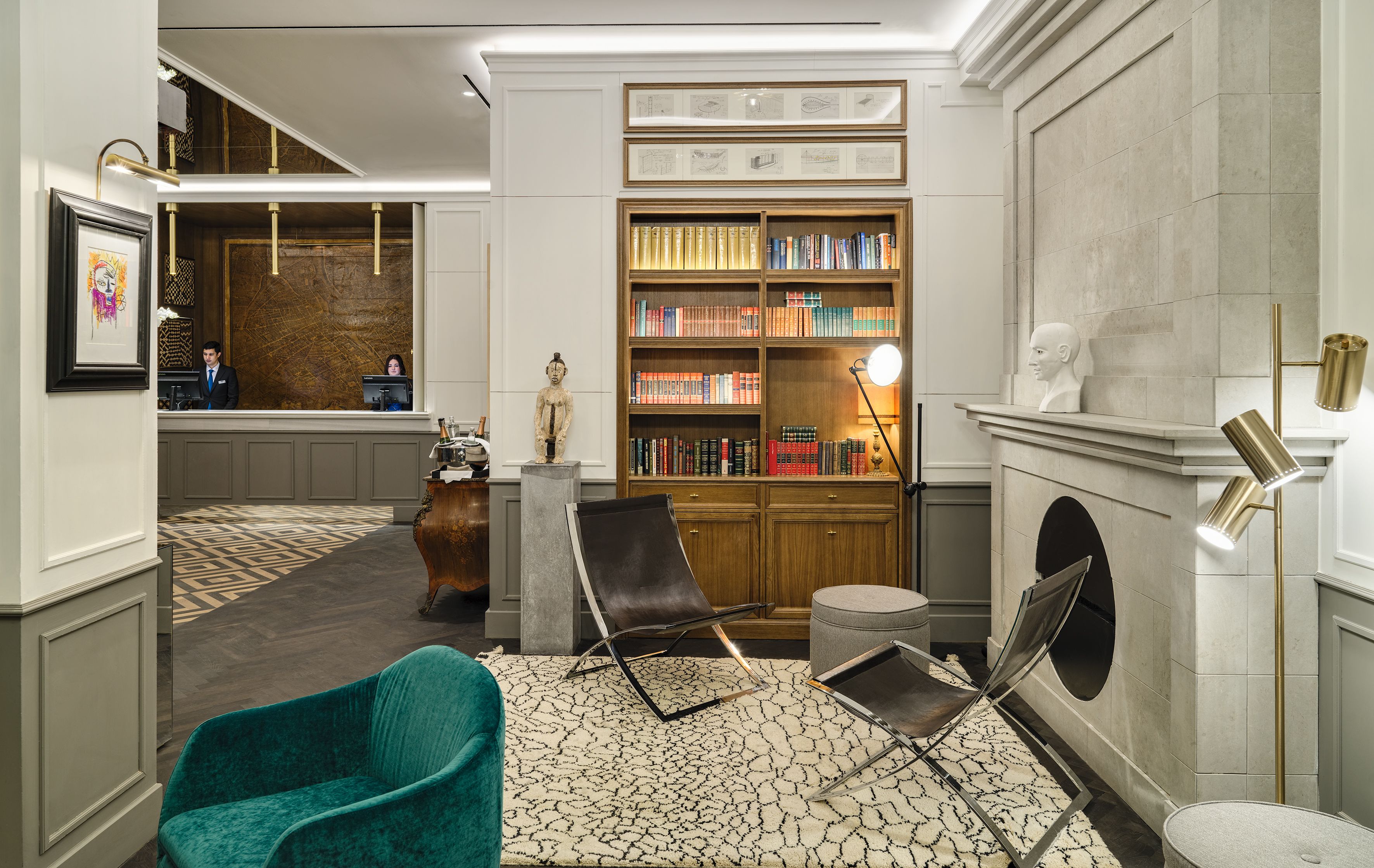 A modern lobby with a reception desk, a large bookshelf, a fireplace, and a lounge area with a teal armchair and a metal-framed chair.