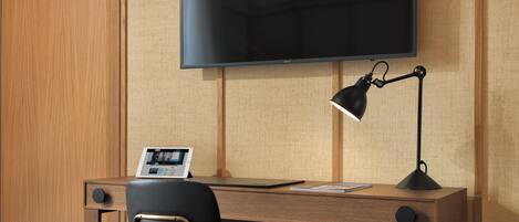 Habitación de hotel con un escritorio de madera, una silla negra, un televisor de pantalla plana montado en la pared y una lámpara de escritorio.
