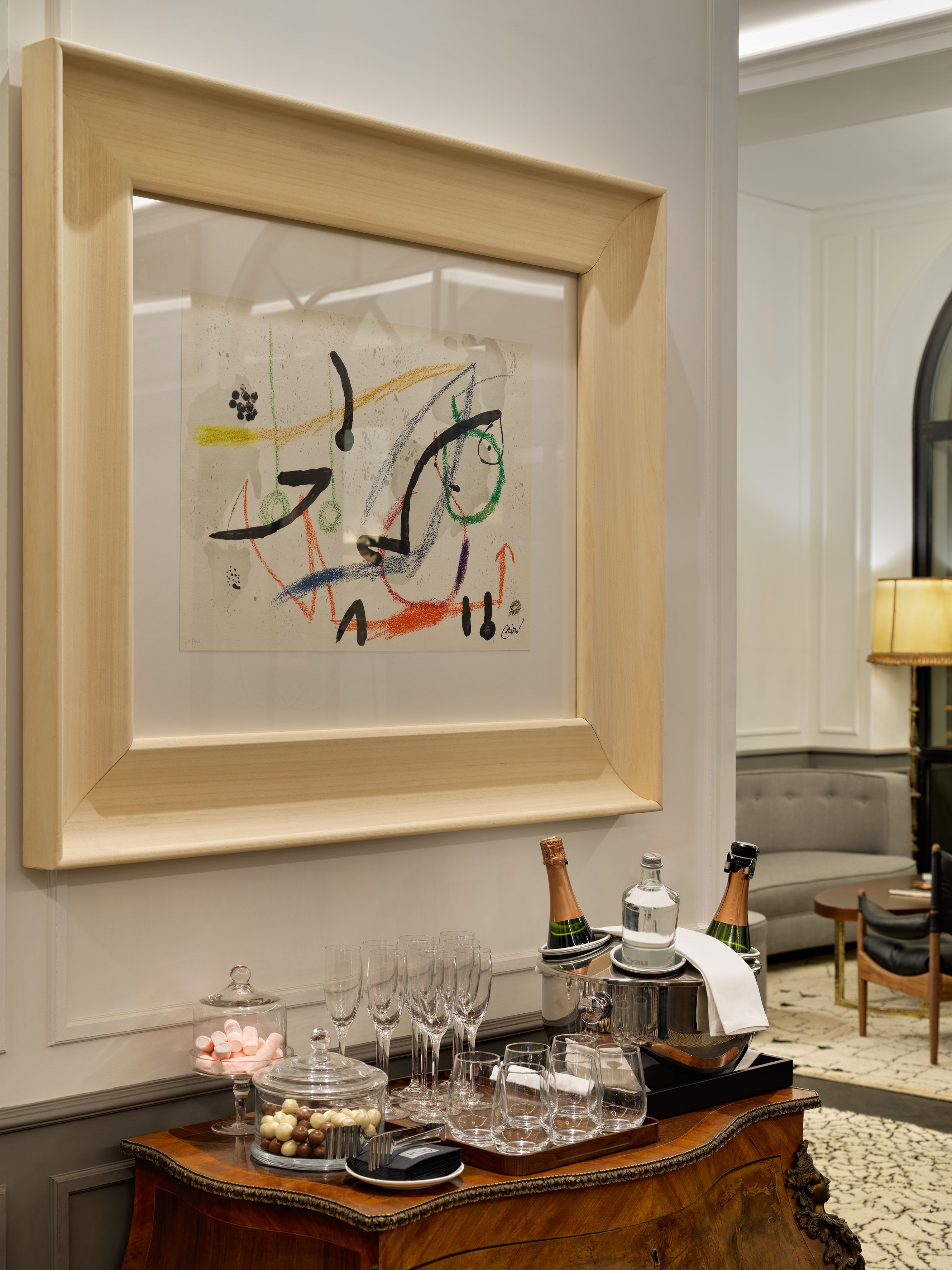 A framed abstract painting on a wall, a wooden sideboard with glasses and bottles, and a grey sofa in the background.
