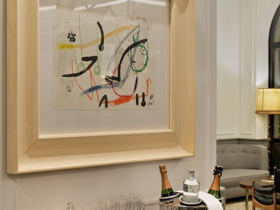 A framed abstract painting on a wall, a wooden sideboard with glasses and bottles, and a grey sofa in the background.
